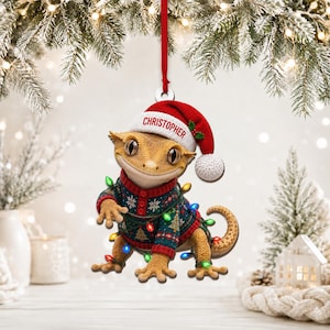 May include: A flat, hanging Christmas ornament featuring a tan gecko wearing a red and white Santa hat with the name Christopher printed on the brim. The gecko wears a dark green and red patterned sweater and is wrapped in colourful string lights.