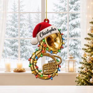 Puede incluir: Un adorno navideño con forma de tuba dorada que lleva un gorro de Papá Noel rojo y blanco con el nombre Christopher impreso en el borde. El instrumento está envuelto en una cadena de luces navideñas multicolores y cuelga de un cordón dorado.