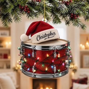 May include: A red glittery snare drum ornament wrapped in multicolored string lights, topped with a red and white Santa hat featuring the name Christopher printed in black script. The ornament hangs from a gold string against a festive background.