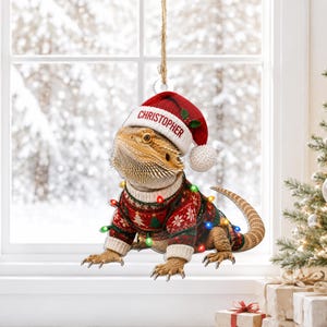 Puede incluir: Un adorno colgante que muestra un dragón barbudo con un gorro de Papá Noel rojo y blanco con el nombre CHRISTOPHER impreso en el borde. El lagarto viste un suéter navideño con estampado rojo y verde adornado con pequeñas luces de colores.