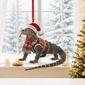 Könnte beinhalten: Ein hängendes Ornament, das einen Waran zeigt, der eine rot-weiße Weihnachtsmütze mit dem Namen CHRISTOPHER auf dem Rand, einen rot-weißen Schal, einen passenden Pullover trägt und mit bunten Miniatur-Weihnachtslichtern umwickelt ist.