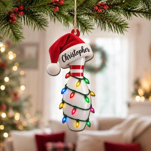 May include: A white bowling pin Christmas ornament featuring a red Santa hat with the name Christopher printed on the brim. The pin is wrapped in a string of colorful, glowing holiday lights in red, green, blue, yellow, and pink hues.