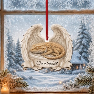 May include: A decorative hanging ornament featuring a tan bearded dragon resting on a cloud, framed by large white feathered wings. A beige banner across the bottom displays the name Christopher in black script, suspended by a red ribbon.