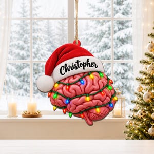 Puede incluir: Un adorno colgante con forma de cerebro humano, que lleva un gorro de Papá Noel rojo y blanco con el nombre Christopher impreso en el borde. El cerebro está envuelto en una cadena de luces navideñas brillantes de colores rojo, verde, azul y amarillo.