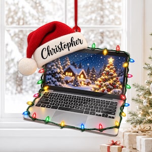 Op de afbeelding: Een hangend ornament in de vorm van een laptop met een rood-witte kerstmuts met de naam Christopher. Het scherm toont een besneeuwd winterlandschap met een verlichte kerstboom, omlijst door een snoer met kleurrijke kerstverlichting.