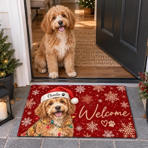 Puede incluir: Un felpudo rectangular rojo con un patrón de copos de nieve dorados y la palabra Welcome en cursiva. La alfombra muestra el gráfico de un perro dorado con un gorro de Papá Noel, el nombre Charlie, una huella de pata y luces navideñas.