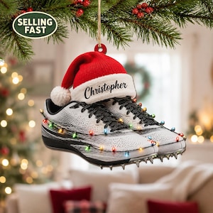 May include: A hanging Christmas tree ornament shaped like a pair of white and black athletic track shoes with spikes on the soles. The shoes are wrapped in miniature colourful Christmas lights and topped with a red and white Santa hat featuring the name Christopher.