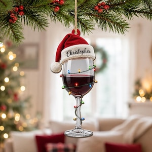 Può includere: Un ornamento piatto da appendere a forma di calice da vino riempito di liquido rosso. Il calice è avvolto da una catena di luci natalizie colorate e sormontato da un cappello da Babbo Natale rosso e bianco con il nome Christopher in nero.