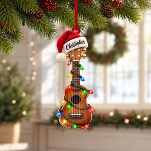 Könnte beinhalten: Ein Weihnachtsbaumschmuck in Form einer hölzernen Ukulele, die eine rote Weihnachtsmütze trägt, auf deren weißem Rand der Name Christopher steht. Das Instrument ist mit einer Lichterkette aus leuchtenden roten, grünen und blauen Lampen umwickelt.