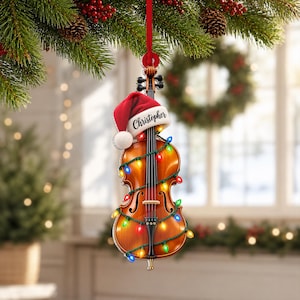 May include: A violin-shaped Christmas ornament with a glossy brown wood finish, wrapped in a string of multicolored LED lights. A red and white Santa hat sits on the top of the violin, featuring the name Christopher printed in black script.