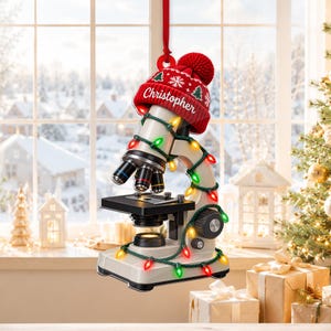 Könnte beinhalten: Ein Mikroskop-Ornament mit einer roten Strickmütze, auf der der Name Christopher in Weiß aufgedruckt ist, verziert mit einer Lichterkette aus bunten Weihnachtsleuchten. Das Mikroskop ist hellgrau mit schwarzen Teilen und hängt an einem roten Band.