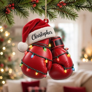 Könnte beinhalten: Ein Weihnachtsbaumschmuck bestehend aus zwei roten Boxhandschuhen, die mit einer Lichterkette aus bunten Miniaturlampen umwickelt sind. Auf den Handschuhen sitzt eine rot-weiße Weihnachtsmütze mit dem Namen Christopher in schwarzer Schrift.