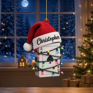 May include: A hanging Christmas ornament shaped like an Ace of Spades playing card, topped with a red and white Santa hat featuring the name Christopher in black script. The card is wrapped in a string of colorful miniature holiday lights.