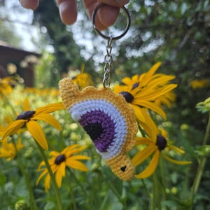 Op de afbeelding: Een gehaakte sleutelhanger met een halfrond oogontwerp met een zwarte pupil, een paarse iris en een wit oogwit, omrand door een gele geschulpte rand. Het textielstuk is bevestigd aan een zilverkleurige metalen ring en ketting.