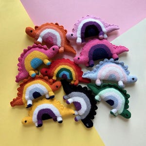 Könnte beinhalten: Eine Ansammlung von zehn kleinen, gehäkelten Dinosaurier-Spielzeugen, die in einer Gruppe angeordnet sind. Jeder Dinosaurier hat ein eigenes Farbmuster mit einem bogenförmigen Design an der Seite, Stacheln am Rücken und schwarzen Perlenaugen.