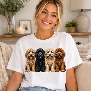 May include: A white short-sleeved t-shirt featuring a graphic print of four fluffy puppies sitting in a row. The dogs are depicted in shades of light brown, black, cream, and dark brown, all with their tongues out in a friendly expression.