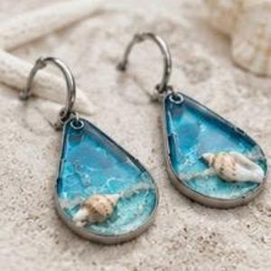 May include: A pair of teardrop-shaped earrings featuring a silver-toned metal frame. The interior contains a blue resin pour mimicking ocean waves with a small, white, spiral seashell embedded at the bottom of each piece.