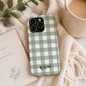 May include: A sage green and white gingham patterned phone case featuring a triple camera cutout at the top and the personalised dark green monogram letters KDM printed in a cursive font near the bottom centre of the rectangular protective cover.