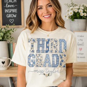 以下が含まれることがあります： クリーム色の半袖Tシャツ。青と白の花柄、ギンガムチェック、水玉模様の大きなブロック体で「THIRD GRADE」とプリントされています。その下には、筆記体で「miss taylor」という名前が書かれています。