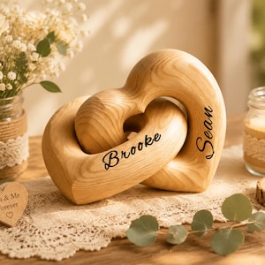 May include: Two interlocking wooden hearts with a natural light brown wood grain finish. The name Brooke is engraved in black script on the front heart, and the name Sean is engraved in black script on the side of the larger heart piece.