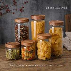Peut inclure: Six bocaux de stockage en verre transparent avec des couvercles en bois ronds, remplis de divers aliments secs dont des grains de poivre, des spaghettis et des pâtes. Le texte 6 SIZES AVAILABLE et 250ML, 350ML, 650ML, 950ML, 1250ML, 1550ML est imprimé.