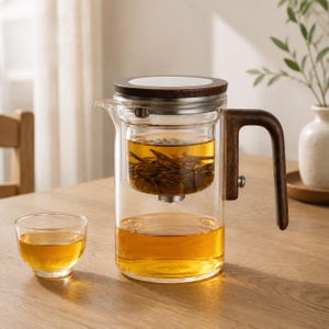 Pode incluir: Um bule de vidro transparente com uma pega de madeira escura e uma tampa a condizer. O bule possui um infusor de vidro interno cheio de folhas de chá, e a base contém chá de cor âmbar. Uma pequena chávena de chá está ao lado.