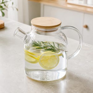 Può includere: Una caraffa in vetro trasparente con coperchio rotondo in legno, manico curvo e beccuccio corto. La caraffa è riempita d'acqua, diverse fette di limone giallo brillante e un rametto di rosmarino verde fresco, creando un semplice contenitore.