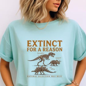 May include: A light blue t-shirt featuring a brown graphic of three dinosaur skeletons. The text reads "EXTINCT FOR A REASON" at the top, with labels "Overconfident," "Unprepared," and "Still Walking" near the skeletons, and "NATURAL SELECTION WAS BUSY" at the bottom.