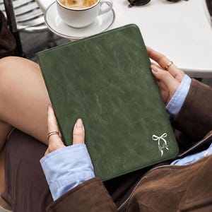 May include: A rectangular, dark olive green tablet case with a smooth, matte texture. The front cover features a small, white, minimalist line-art illustration of a tied ribbon bow positioned in the bottom right corner of the protective sleeve.