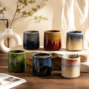 Puede incluir: Una colección de seis tazas cilíndricas de cerámica con un acabado de esmalte reactivo y patrones de colores que gotean. Las tazas muestran varias combinaciones de colores en verde, azul, negro, marrón y crema, con una textura lisa y brillante.