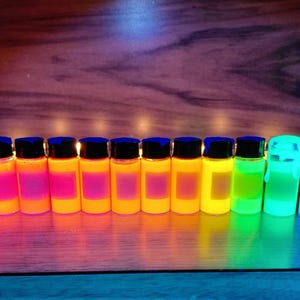 May include: A row of fourteen small, clear glass vials filled with vibrant, fluorescent-colored liquids, each topped with a black cap. The liquids glow in shades of pink, orange, yellow, green, and blue, arranged in a spectrum-like sequence.