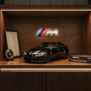 Könnte beinhalten: Ein schwarzes BMW M3 Modellauto auf einem Holzregal. Hinter dem Auto befindet sich ein metallisches BMW M Logo. Links steht eine gerahmte technische Zeichnung und eine Bremsscheibe. Rechts liegen zwei Bücher mit der Aufschrift BMW M und THE ULTIMATE DRIVING MACHINE.