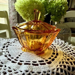 May include: A translucent amber glass candy dish with a matching lid featuring a faceted, geometric design and a central finial handle. The bowl has a scalloped rim and rests on three small feet, showcasing a vibrant orange hue and reflective surface.