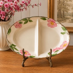 May include: An oval-shaped, cream-colored ceramic serving dish with a central divider. The rim features a painted floral pattern of pink flowers and green leaves. The dish is displayed on a small, dark-toned metal stand for upright presentation.