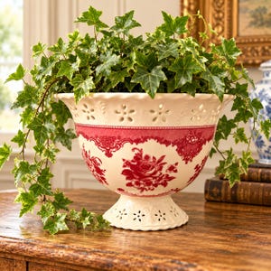 May include: A cream-colored ceramic pedestal bowl featuring a red floral pattern and a scalloped rim with cut-out flower details. The bowl is filled with a lush green ivy plant that cascades over the sides, creating a vibrant contrast.
