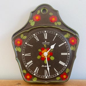 May include: A dark brown ceramic wall clock with a scalloped edge and a hanging hole. The clock face features white Roman numerals, white hands, and a central floral motif in red, orange, and green. Text reads Junghans Quartz and Handgealt.