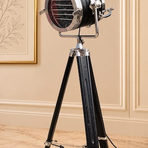 May include: A floor lamp featuring a black cylindrical spotlight head with a chrome rim and front grille, mounted on a black wooden tripod stand with chrome hardware and a thin metal chain connecting the three legs at the base.