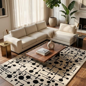 Op de afbeelding: Een rechthoekig vloerkleed met een crèmekleurige basis en een herhalend zwart geometrisch patroon van cirkels en lijnen. Het kleed ligt op een houten vloer onder een houten salontafel, een witte vaas en een stapel boeken.