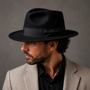 May include: A black fedora hat featuring a structured crown and a wide brim, accented with a matching black grosgrain ribbon band around the base. The hat is worn over a black collared shirt and a light beige textured blazer with notched lapels.