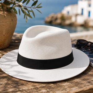 May include: A white fedora hat with a wide, flat brim and a pinched crown, featuring a solid black grosgrain ribbon band encircling the base of the crown. The hat is constructed from a finely woven material with a consistent, tight texture.