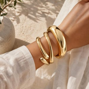 Könnte beinhalten: Ein poliertes, goldfarbenes Armband mit einem dicken, abgerundeten Spiraldesign, das sich um das Handgelenk legt. Das Metall hat eine glatte, reflektierende Oberfläche mit einem Hochglanz-Finish, was ein fließendes und skulpturales Aussehen erzeugt.