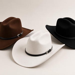 Peut inclure: Trois chapeaux de cowboy en marron, blanc et noir. Chaque chapeau présente une calotte plissée, un large bord incurvé et une fine bande noire autour de la base avec une petite boucle argentée. Les chapeaux sont disposés en groupe sur une surface neutre.
