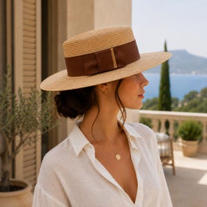 May include: A straw boater hat with a flat crown and a wide brim, featuring a dark brown satin ribbon band tied in a bow with a thin gold-tone metal accent piece. The hat is constructed from woven natural tan straw with a structured, rigid shape.