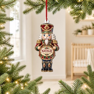 May include: A nutcracker ornament hanging from a red ribbon, featuring a soldier in a red, gold, and dark blue uniform with a white beard. The figure holds a drum with the name NATALIE and the year 2026 printed on the front surface.