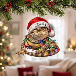 May include: A hanging ornament featuring a bearded dragon wearing a red and white Santa hat with the name Bentley printed on the brim. The lizard is wrapped in a string of colourful Christmas lights with red, green, blue, and yellow bulbs.