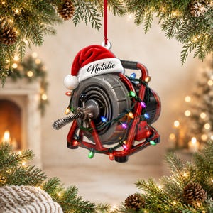 May include: A red drain cleaning machine ornament featuring a Santa hat with the name Natalie printed on the white trim. The machine is wrapped in colourful Christmas lights and hangs from a red ribbon against a festive holiday background.