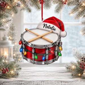 Puede incluir: Un adorno navideño de tambor rojo con parche blanco y dos baquetas de madera cruzadas encima. Un gorro de Papá Noel rojo con el nombre Natalie impreso en negro descansa sobre el tambor, rodeado por una guirnalda de luces navideñas de colores.