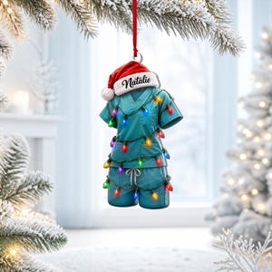 Puede incluir: Un adorno con forma de casaca y pantalón de pijama médico en color verde azulado, colgado de una cinta roja. El conjunto está envuelto en una guirnalda de luces navideñas de colores y rematado con un gorro de Papá Noel con el nombre Natalie.