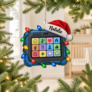 May include: A blue tablet-shaped Christmas ornament featuring a red Santa hat with the name Natalie printed in black script. The device screen displays a grid of twelve colorful icons, and the frame is wrapped in a string of multicolored lights.