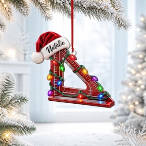 May include: A red triangular drafting square ornament featuring a white Santa hat with the name Natalie printed in black script. The tool is wrapped in a string of multicolored Christmas lights and hangs from a thin red ribbon.